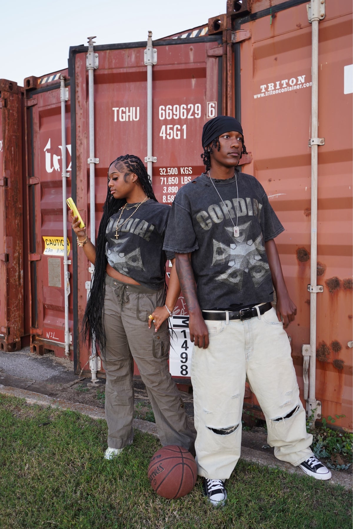 Two models wearing charcoal Cordial tees beside shipping containers